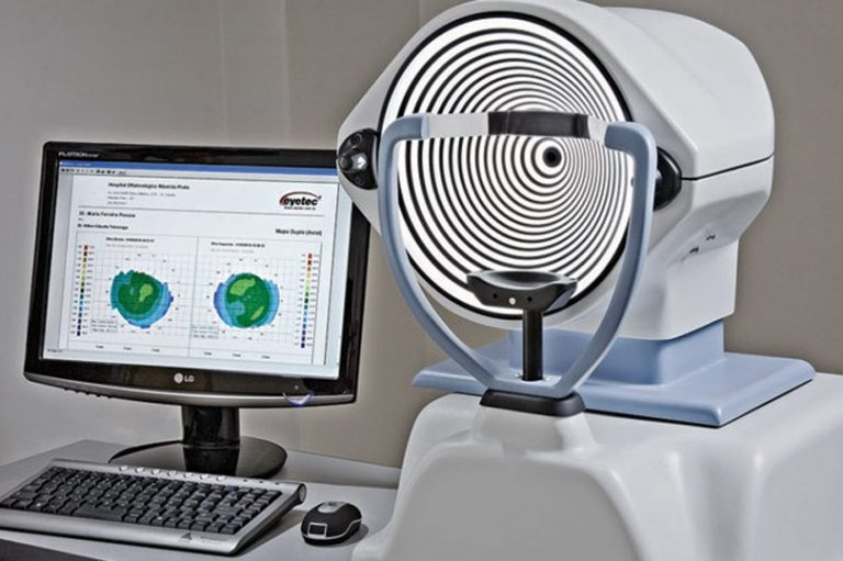Imagem de aparelho utilizado para realizar o exame de topografia corneana ao lado de um computador mostrando resultados de exame