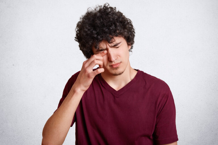 Tired teenager with crisp hair, rubs eyes as wants to sleep, has bad eyesight, dressed in red casual t shirt, isolated over white concrete wall. People, tiredness and health problems concept